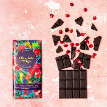 Tablete de chocolate preto biológico e vegan Chocolate and Love, 70% cacau com romã, partida em pedaços sobre superfície rosa, acompanhada por sementes de romã.