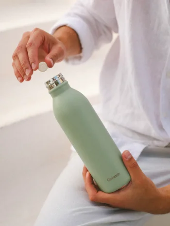 Pessoa segura garrafa térmica Qwetch de 600ml em aço inox, cor verde tília, e coloca uma pastilha de solid drink, para dissolver na água, dentro da garrafa.