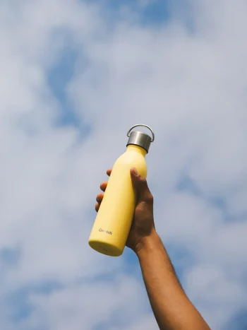 Mão segura garrafa térmica Qwetch de 600ml em aço inox, cor limão, com tampa metálica e pega, contra céu nublado.