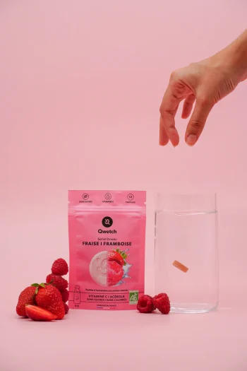 Mão a colocar uma pastilha num copo de água ao lado de embalagem rosa de Solid Drink Bio sabor morango e framboesa da Qwetch, com morangos e framboesas frescas. Compra já online!