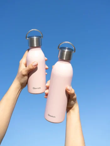 Duas mãos seguram garrafas térmicas Qwetch em aço inox, cor rosa pastel, contra um céu azul. Garrafa de 350ml à esquerda e garrafa de 600ml à direita.