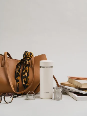 Garrafa térmica Qwetch de 400ml na cor areia e o infusor, colocada sobre superfície com mala castanha, lenço com padrão leopardo, óculos e livros empilhados.