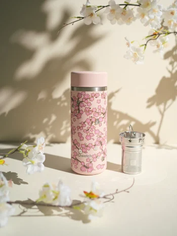 Garrafa de chá térmica Qwetch Flowers rosa pastel, 400ml, com padrão de flores de cerejeira (sakura) em tons de rosa e branco, acompanhada de infusor metálico, sobre fundo com sombras florais.