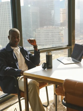 Pessoa sentada à mesa junto a janela com vista urbana, com garrafa térmica preta sobre a mesa, em ambiente de escritório moderno.