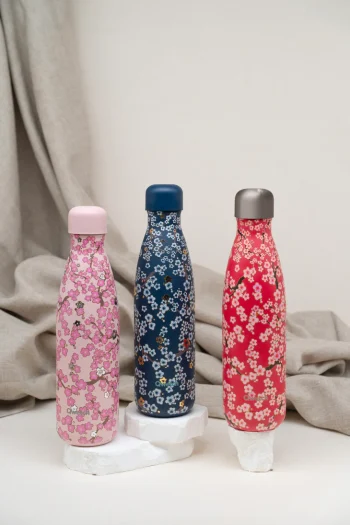 Três garrafas térmicas reutilizáveis Qwetch Originals Flowers de 500ml com padrões florais: rosa, azul escuro e vermelha, dispostas sobre pedestais brancos com fundo neutro e tecido drapeado.