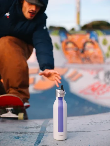 Garrafa térmica Qwetch de 600ml com tampa sport, cor cinza e faixa violeta, colocada sobre superfície em ambiente outdoor com skate ao fundo.