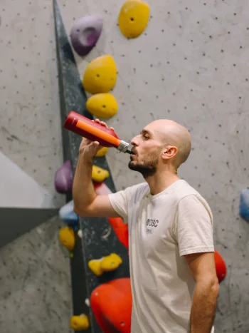 Pessoa a beber de uma garrafa térmica Qwetch 600ml com tampa sport, cor vermelho grenat e faixa laranja, em ambiente de escalada indoor.